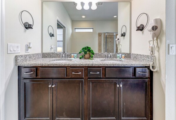A bathroom with a granite countertop, dark wooden cabinets, a large mirror, and a hairdryer mounted on the wall.