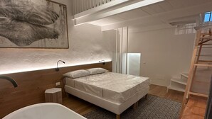Moderni makuuhuone, jossa on suuri valkoinen kylpyamme, valkoisella patjalla varustettu sänky, puulattia ja korkea katto, jossa on valkoinen kaide.