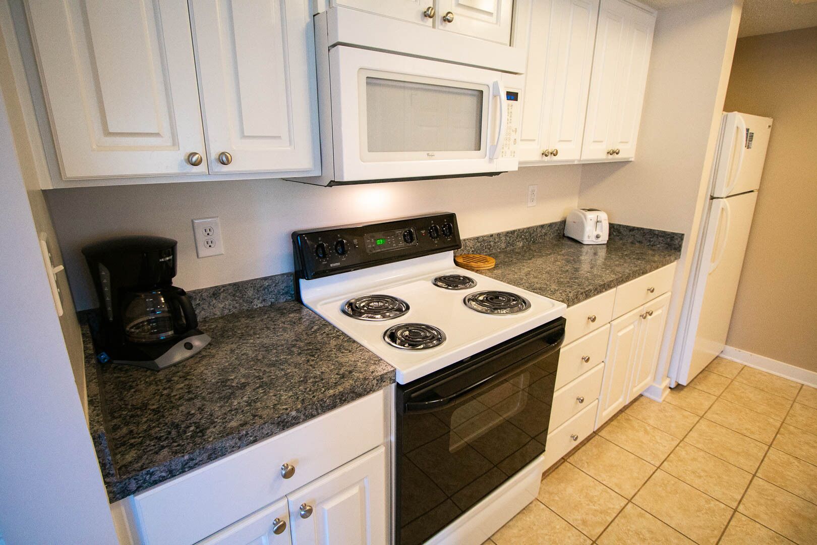 Una cocina con gabinetes blancos, estufa, microondas, cafetera y refrigerador.