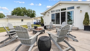 Eine Terrasse mit Adirondack-Stühlen und einem Feuerkorb, ein weißes Haus mit großen Fenstern und ein weißer Zaun im Hintergrund.