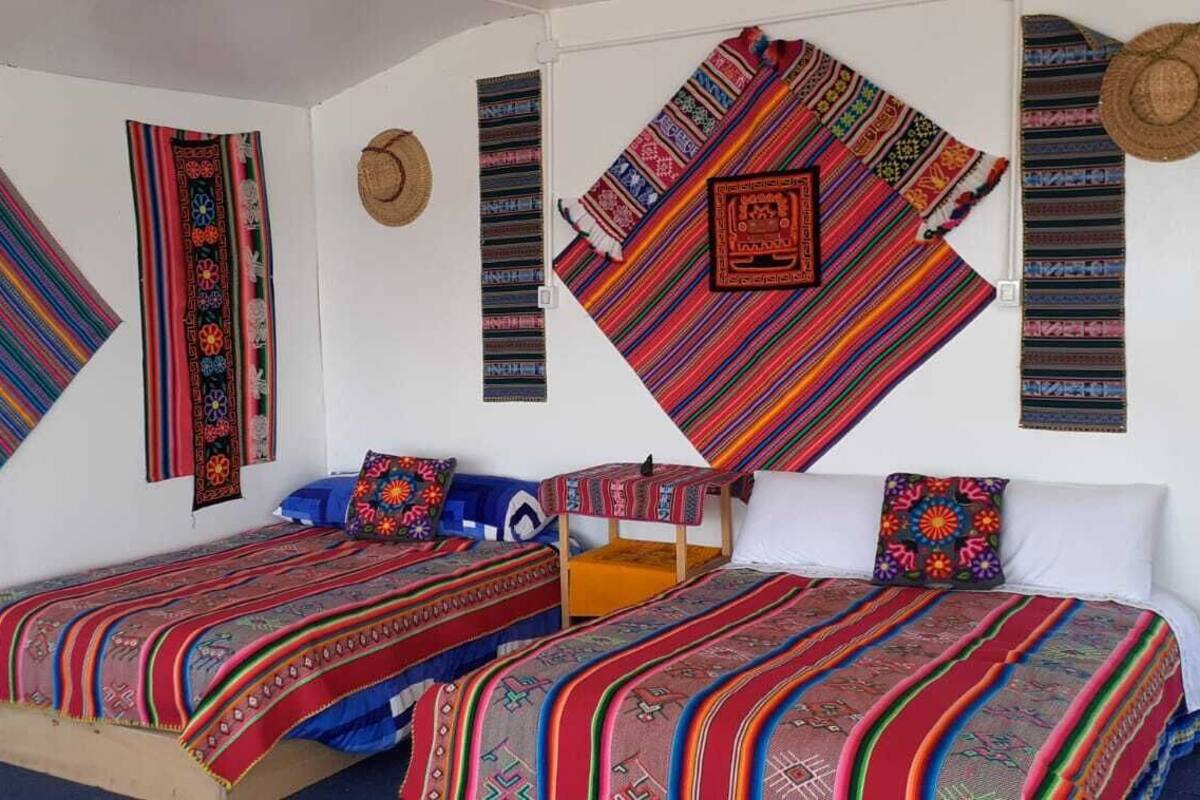 Una habitación con dos camas, cada una con mantas de colores, y una pared decorada con tejidos y sombreros.