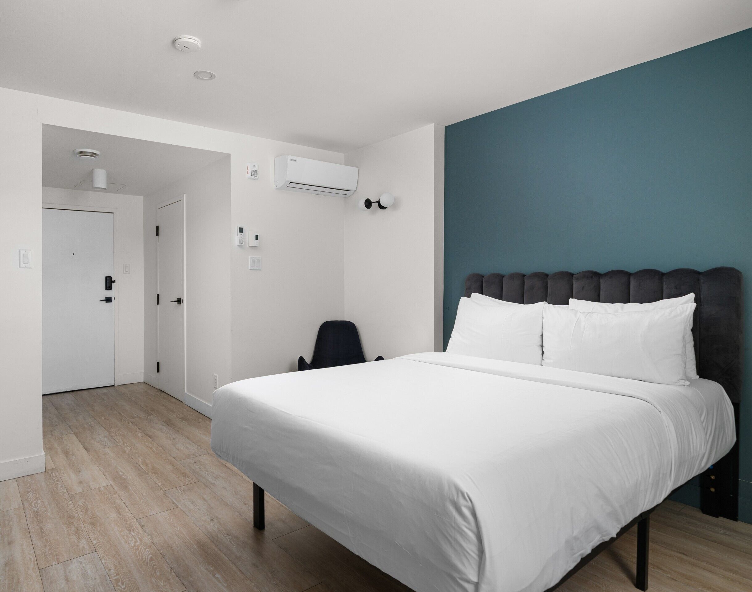 A hotel room with a bed, a chair, a door, and an air conditioning unit.