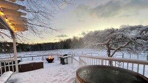 Een sneeuwbedekt terras met een hottub, vuurkorf en lichtjes.