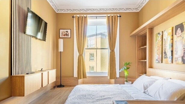 Ein Hotelzimmer mit einem Bett, einem Nachttisch, einem an der Wand montierten Fernseher, einem Fenster mit Vorhängen und einer Pflanze auf dem Fensterbrett.