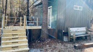 A wooden deck leading to a cabin with a metal door and a bench outside. - House - The Fais Do Do (Hattiesburg)