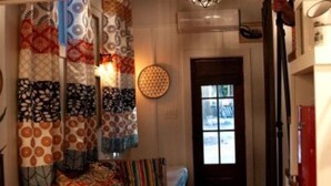 A cozy room with a bed, colorful curtains, a round wall lamp, and a door leading to another room. - Tiny House - Shangri La (Hattiesburg)