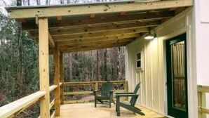 Una terraza de madera con dos sillas y una mesa, rodeada de un bosque.
