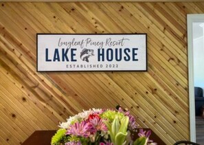 Schild für das Lake House des Longleaf Piney Resort, gegründet 2022, mit Holzvertäfelung im Hintergrund und einer Blumendekoration darunter.