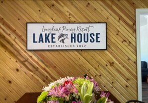Naamplaatje voor Lake House van Longleaf Piney Resort, opgericht in 2022, met een houten achtergrond en een bloemstuk eronder.