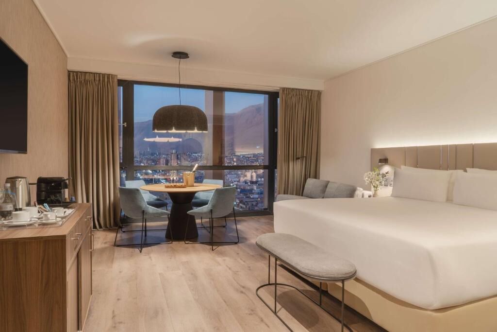 A hotel room with a large bed, a dining area with a table and chairs, a TV, and a city view through the window.