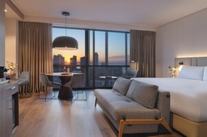Habitación de hotel moderna con una cama grande, un sofá, un escritorio y una mesa de comedor. Se ve una vista de la ciudad a través de ventanas amplias.