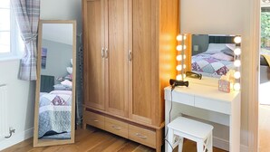 Un dormitorio con un armario de madera, una tocador con luces, un espejo, una cama con colcha y una ventana con cortinas.