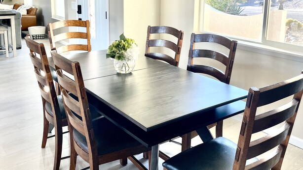 A dining area with a rectangular table surrounded by six wooden chairs with metal legs. There is a vase with flowers on the table, and the room has large windows providing a view of the outdoors.