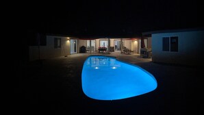 A swimming pool with a blue light, surrounded by a covered patio with tables and chairs.