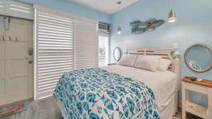 Een slaapkamer met een bed, een raam met zonwering, een wandspiegel en een klein tafeltje met een lamp.