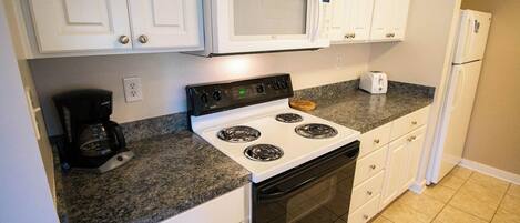 Una cocina con gabinetes blancos, estufa, microondas, cafetera y refrigerador.