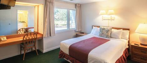 A hotel room with a bed, a desk with a telephone, a chair, a window with curtains, and a nightstand with a lamp.