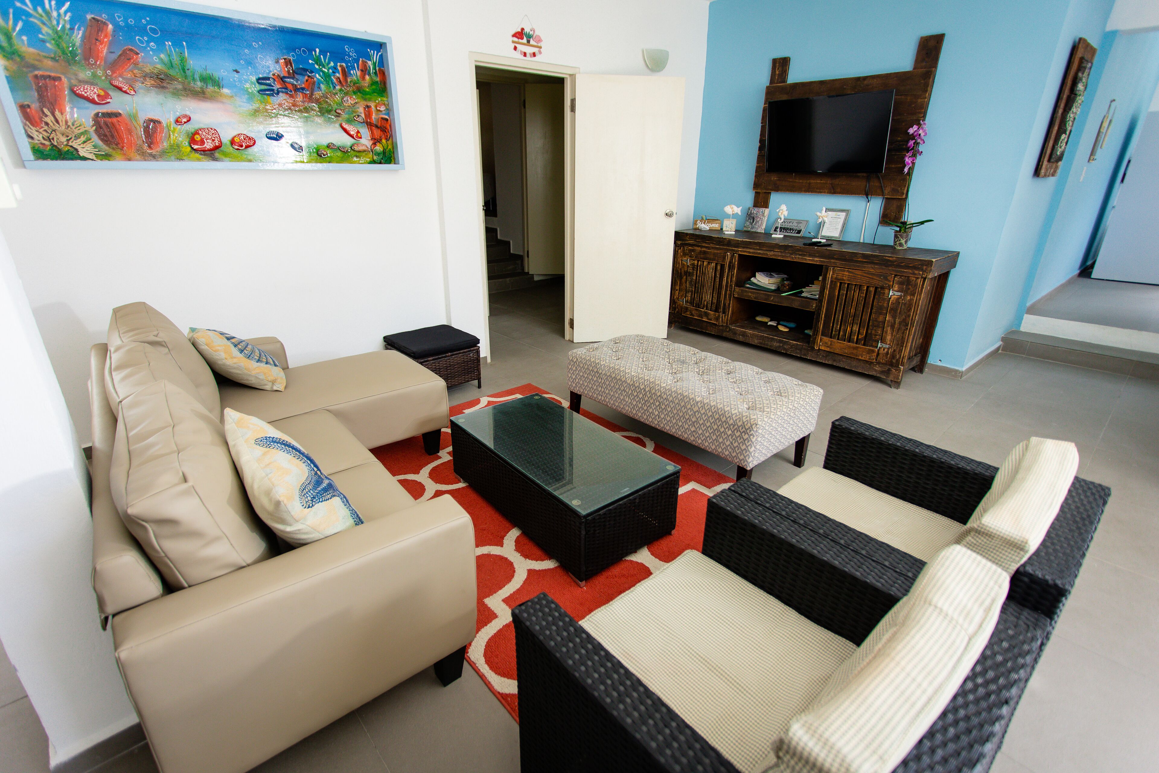 Un salon comprenant un canapé, un pouf et une table basse, un téléviseur posé sur un meuble en bois, et un tableau accroché au mur.