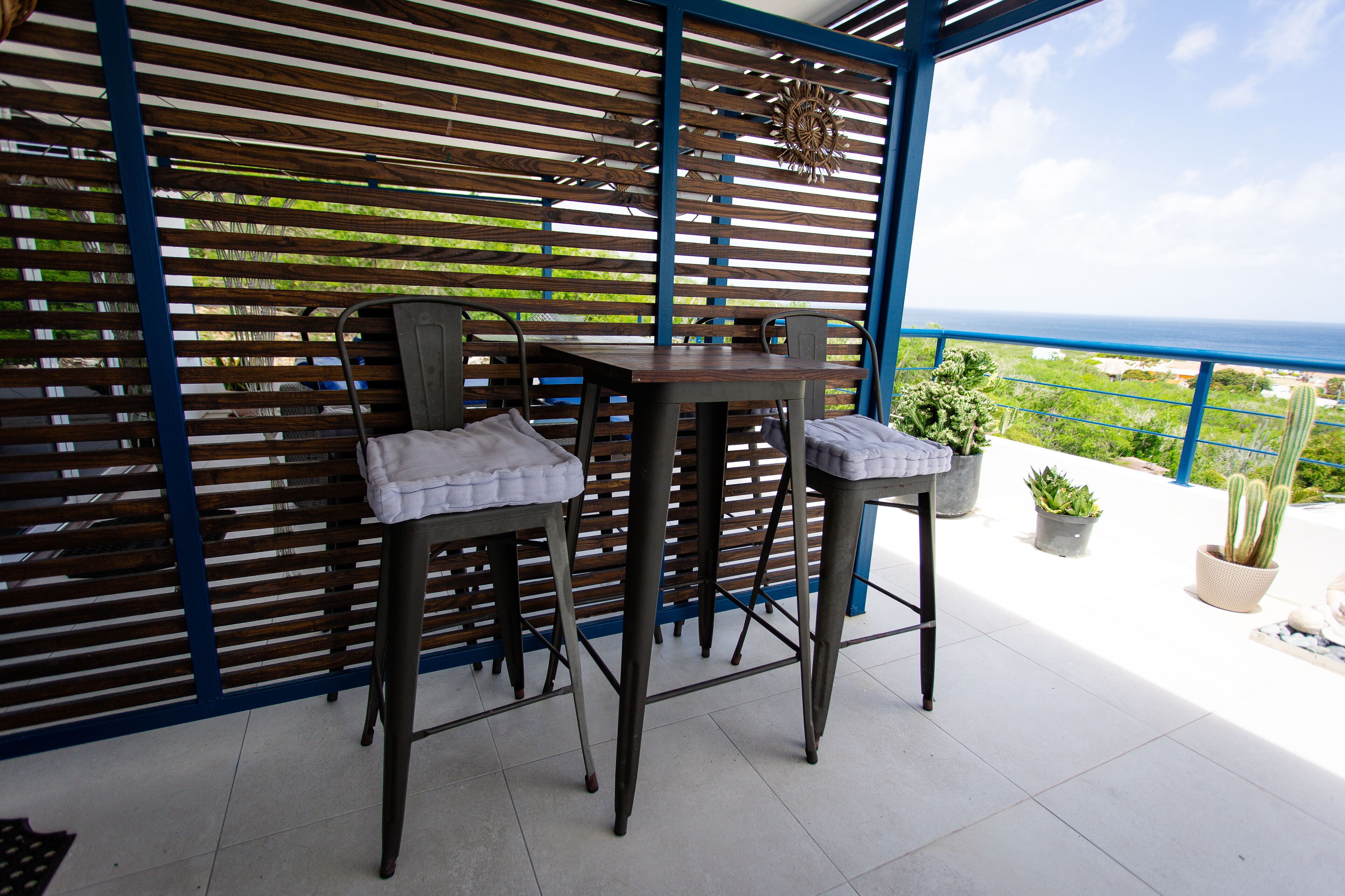 Una terraza con dos taburetes de bar y una mesa, un muro de listones de madera y plantas en macetas.