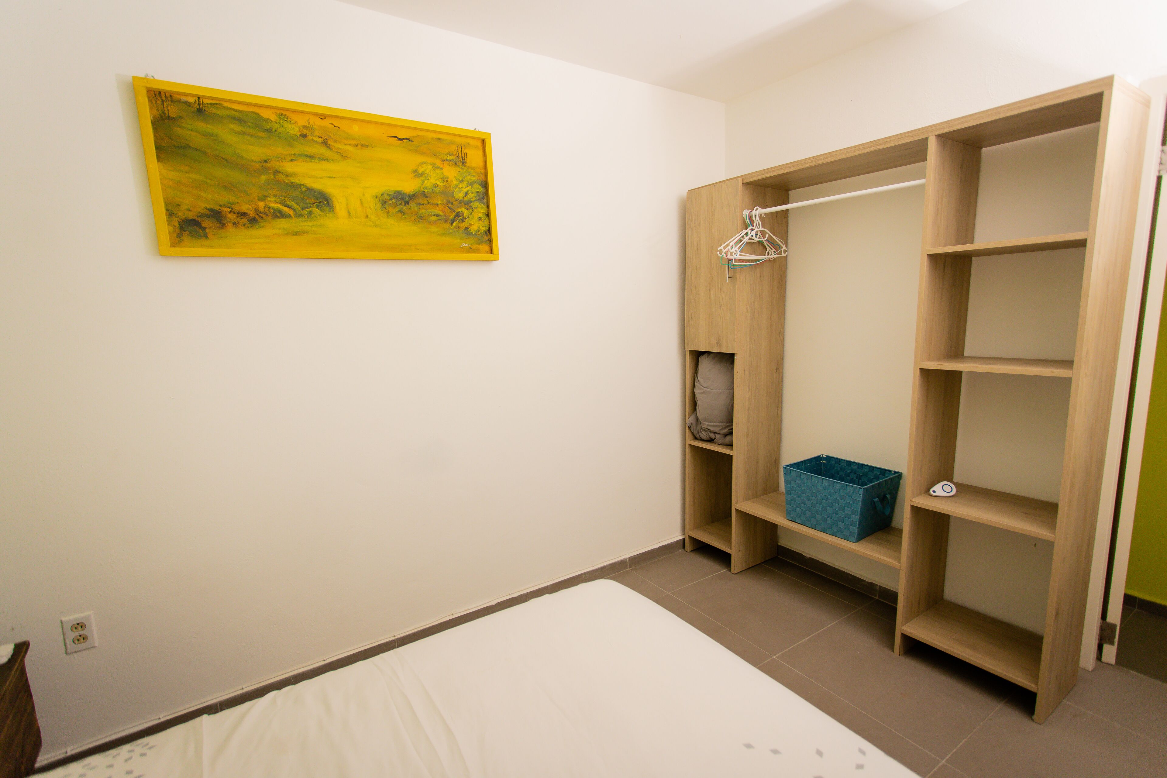 A bedroom with a bed, a wooden wardrobe, a blue storage box, and a yellow painting on the wall.