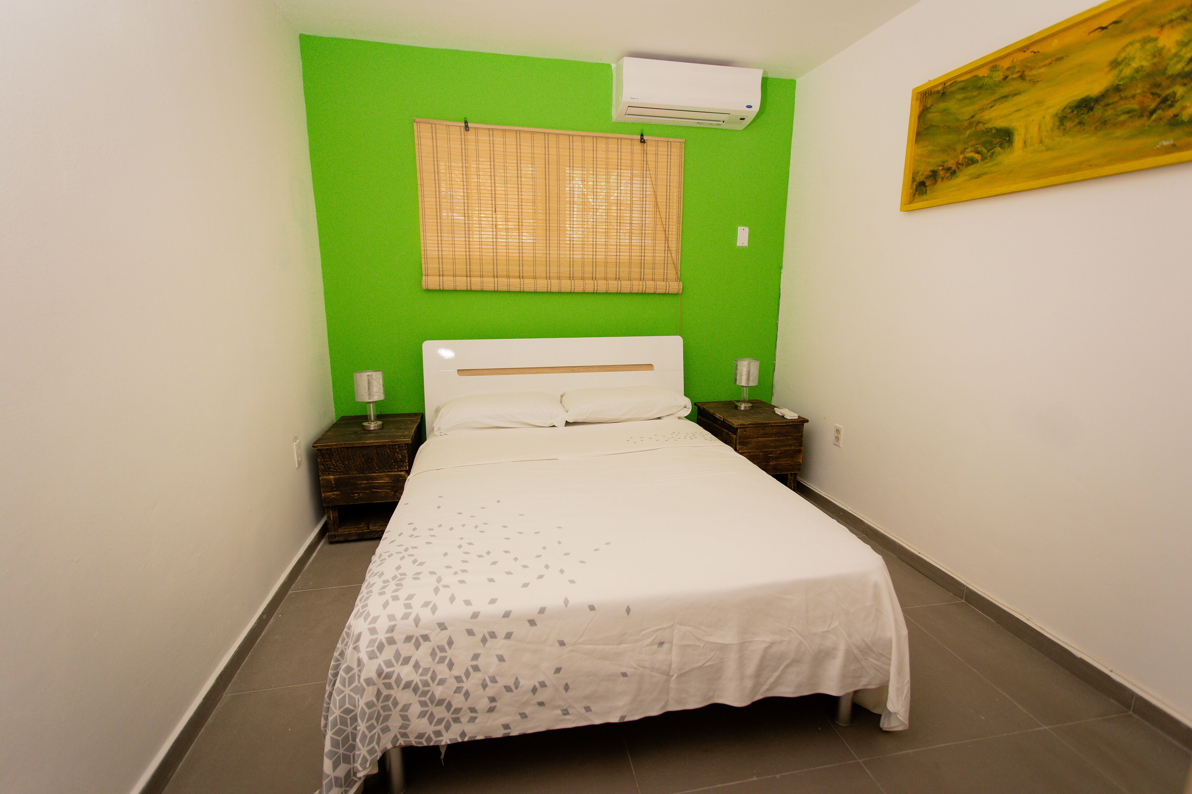 A small, neatly arranged bedroom with a single bed, a bedside table, a nightstand, a wall-mounted air conditioner, and a window with blinds.