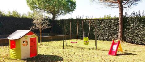 Un parque infantil con columpios, una casita de juegos y un árbol.