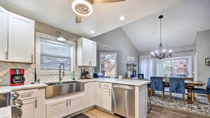 A modern kitchen with white cabinets, a stainless steel sink, and a dishwasher. There is a dining area with a wooden table and chairs. - Lakefront Rogers Getaway w/ Hot Tub & Deck! (Rogers)