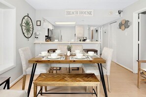 A dining area with a wooden table set for four, surrounded by upholstered chairs. A bench is placed in front of the table. The room has a neutral color palette with white walls and a ceiling light. - OCEAN FRONT Gem w/Pools, Movie Theater, Game Room, SunsetCove at SeaWatch (Ocean City)