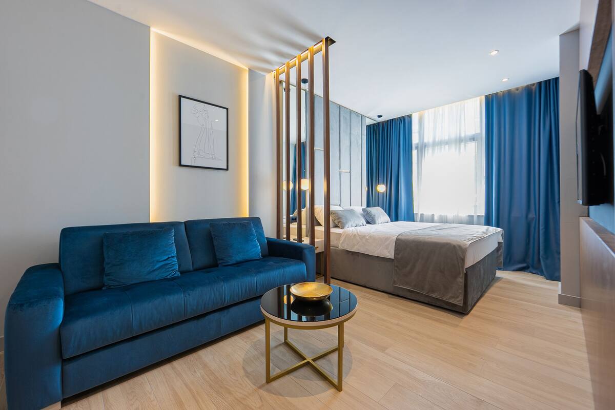Una habitación de hotel moderna con un sofá azul, una mesa de centro redonda, una cama con edredón gris y un ventanal con cortinas azules.