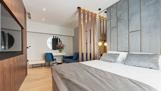 A modern hotel room with a large bed, a desk with a chair, a round mirror, and a decorative wall panel. - Caldo Luxury Rooms (Split)