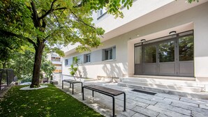 A modern building with a stone-paved courtyard, benches, and a tree providing shade. - Caldo Luxury Rooms (Split)