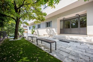 Un edificio moderno con un cortile lastricato in pietra, panchine e un albero che offre ombra.