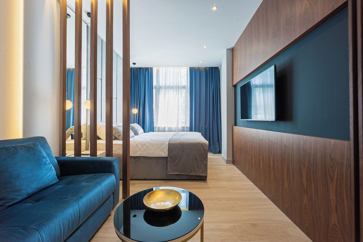 Una habitación de hotel moderna con un sofá azul, una cama con edredón blanco, una mesa de centro negra redonda con un cuenco y un televisor montado en la pared.