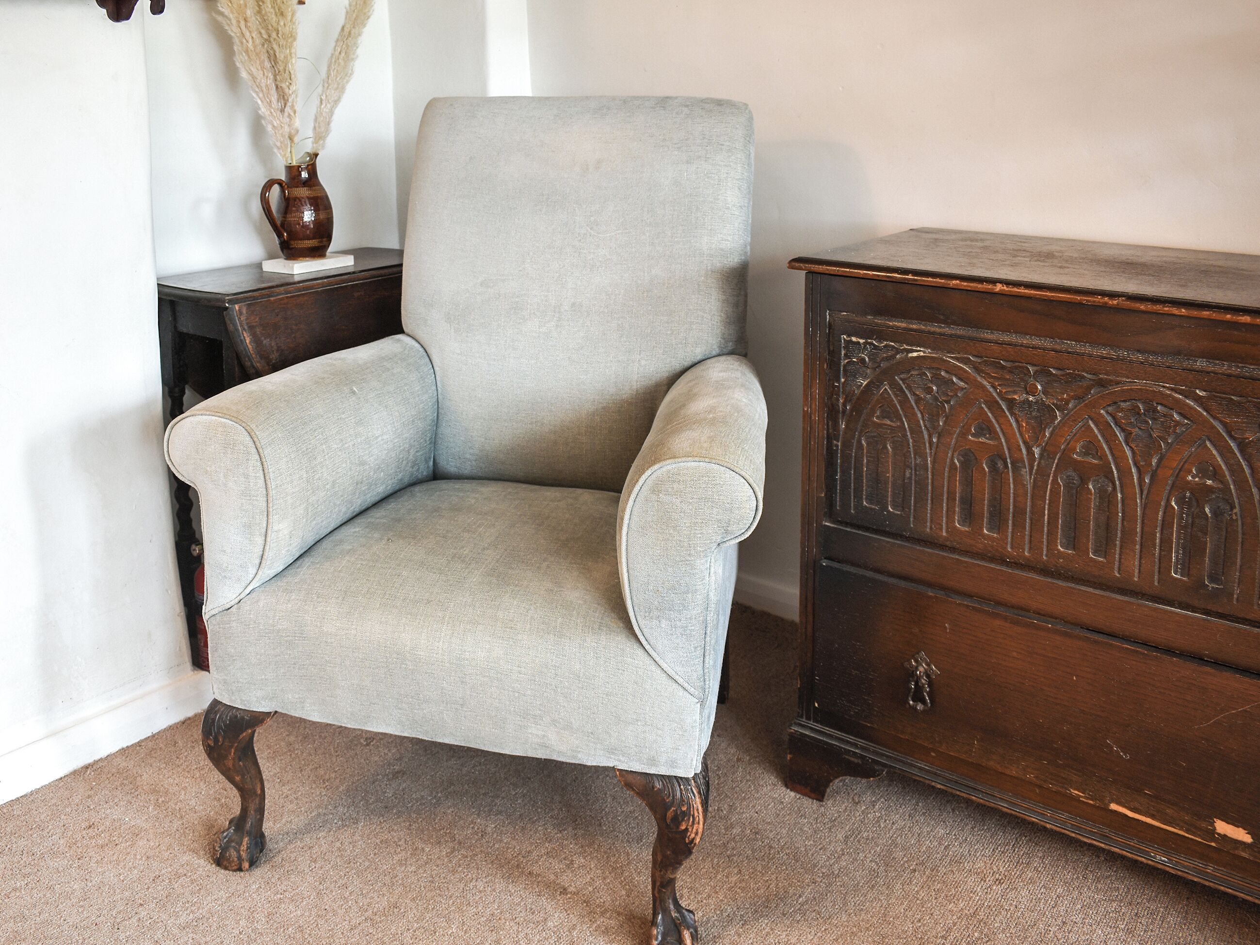 Un sillón tapizado de gris claro con patas de madera, ubicado junto a un aparador de madera oscura con detalles tallados.