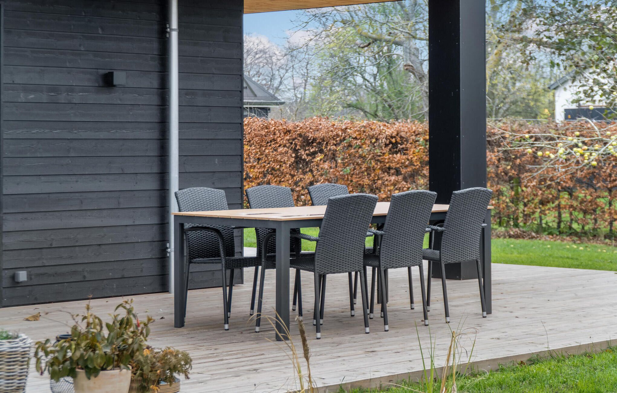A covered outdoor dining area with a table and chairs, a wooden deck, and a hedge in the background.