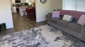 A living room with a gray couch, a patterned rug, and a kitchen in the background. - Downtown Taber Street Railroad 🚂 House - Great Location!! (Williams)