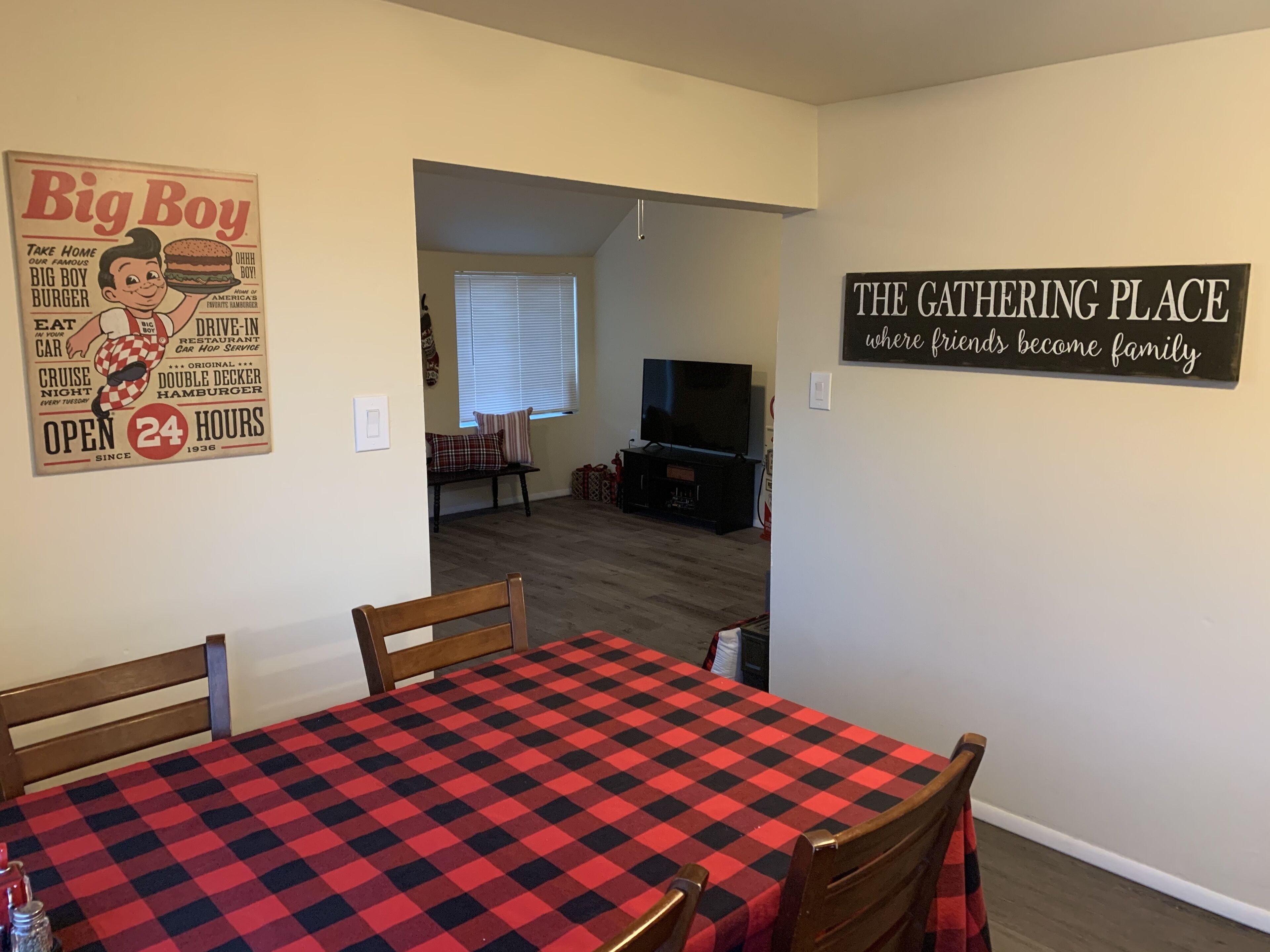 Et spiseområde med et ternet borddække, et fjernsyn i baggrunden, og et skilt på væggen med teksten "THE GATHERING PLACE hvor venner bliver familie".