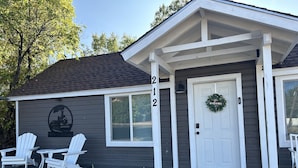 A building with a dark gray exterior, white trim, and a white door with a wreath. There are two white Adirondack chairs and a small table outside. - Downtown Taber Street Railroad 🚂 House - Great Location!! (Williams)