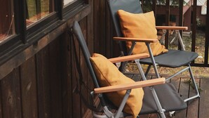 Twee opklapbare stoelen met kussens op een houten terras, naast een raam met uitzicht op bomen en een gebouw.