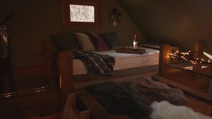 Een gezellig zolderkamer met een hoogslaper, een kleine tafel met een fles en glazen, en een raam met uitzicht op kale bomen.