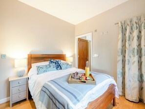 A bedroom with a wooden bed, a bedside table with a lamp, a tray with fruits and a bottle, and a floral curtain. - 1 bedroom accommodation in Diss (Diss)