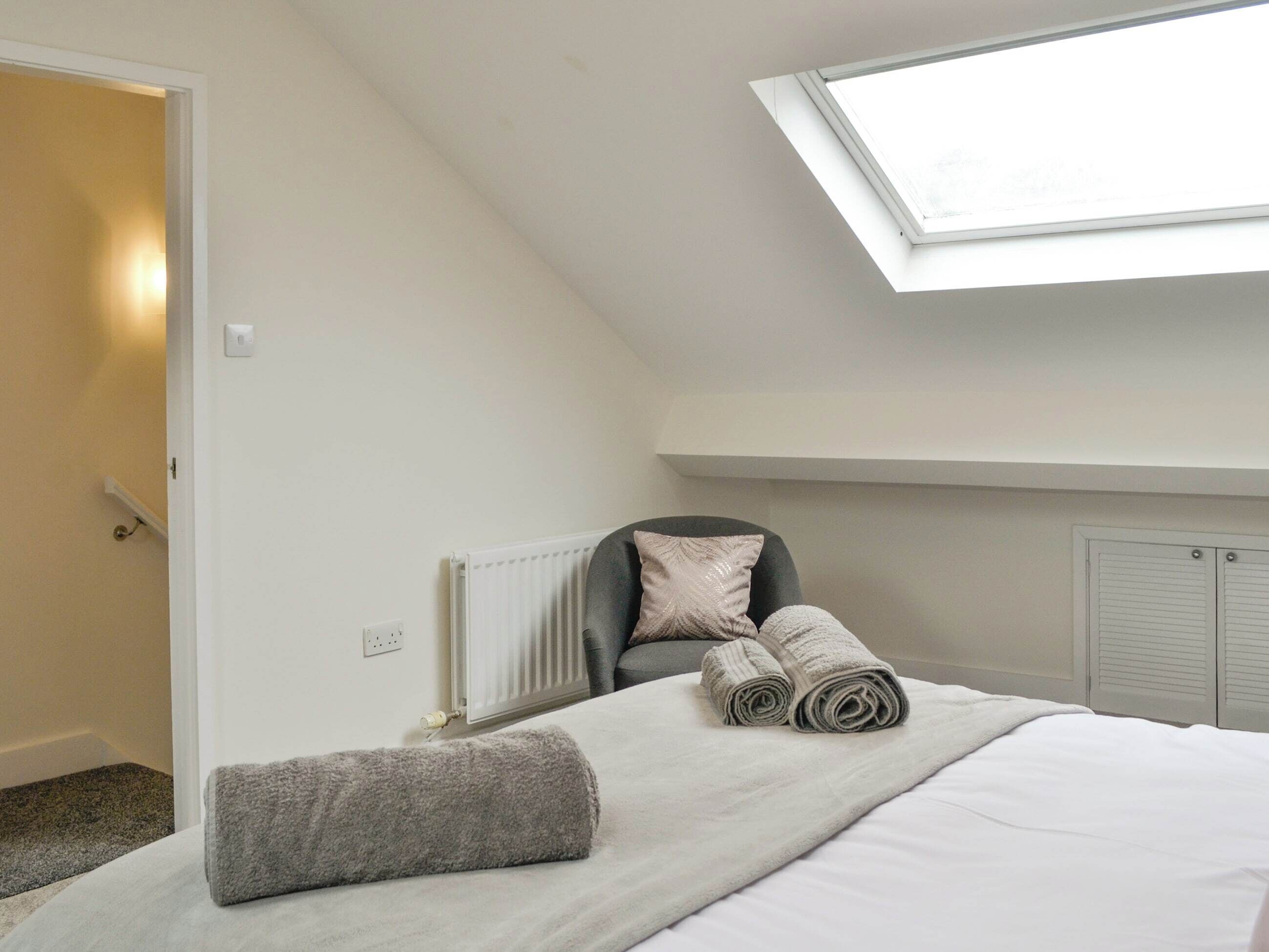 A bedroom with a skylight, a radiator, a bed with rolled towels, and a grey chair.