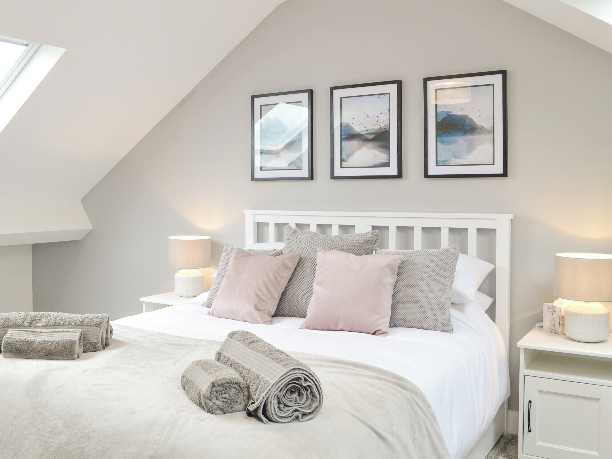 A bedroom with a sloped ceiling, a white bed with multiple pillows, two bedside tables with lamps, and three framed pictures on the wall.