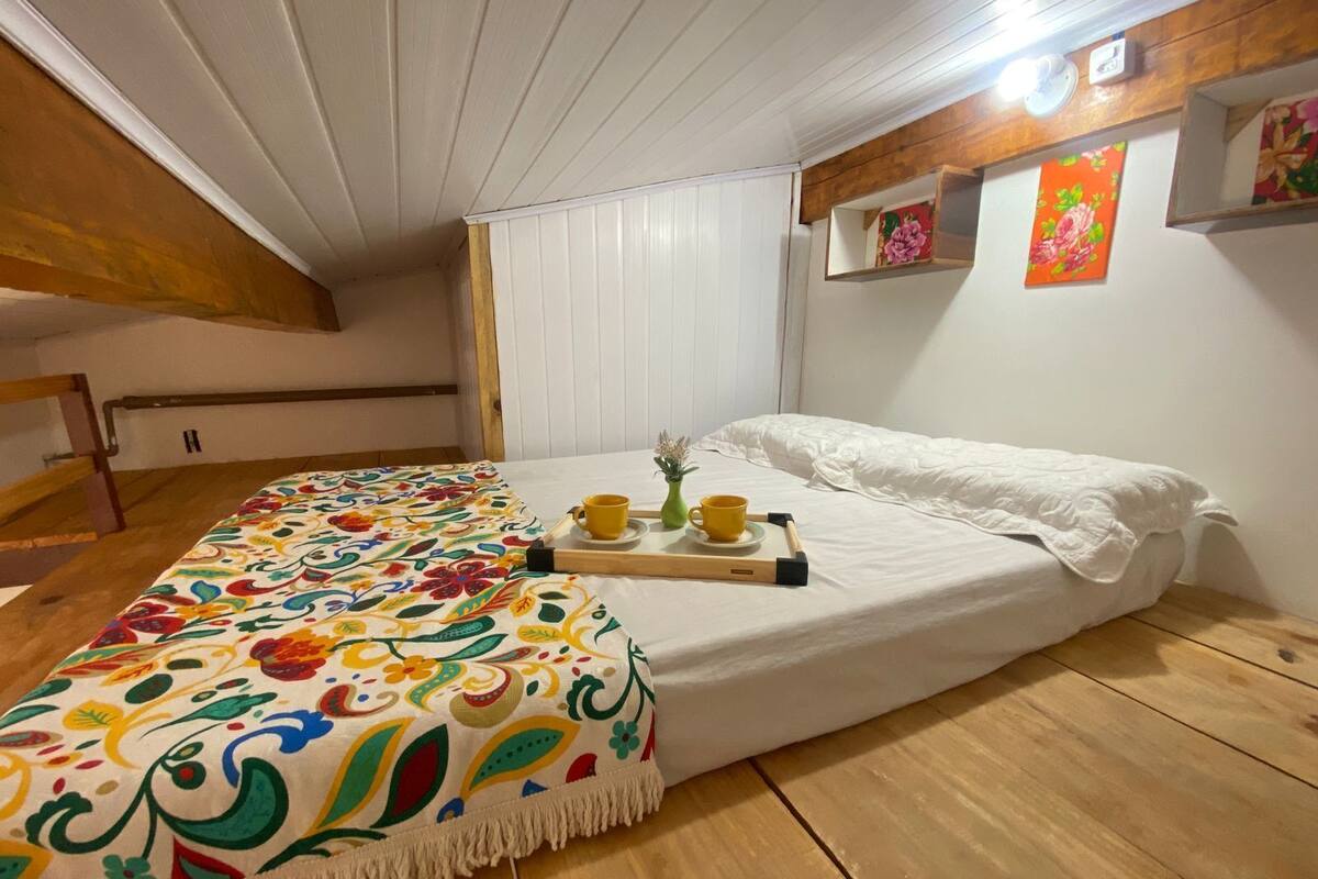 A single bed with a colorful blanket, a bedside table with a cup and saucer, and a small plant.