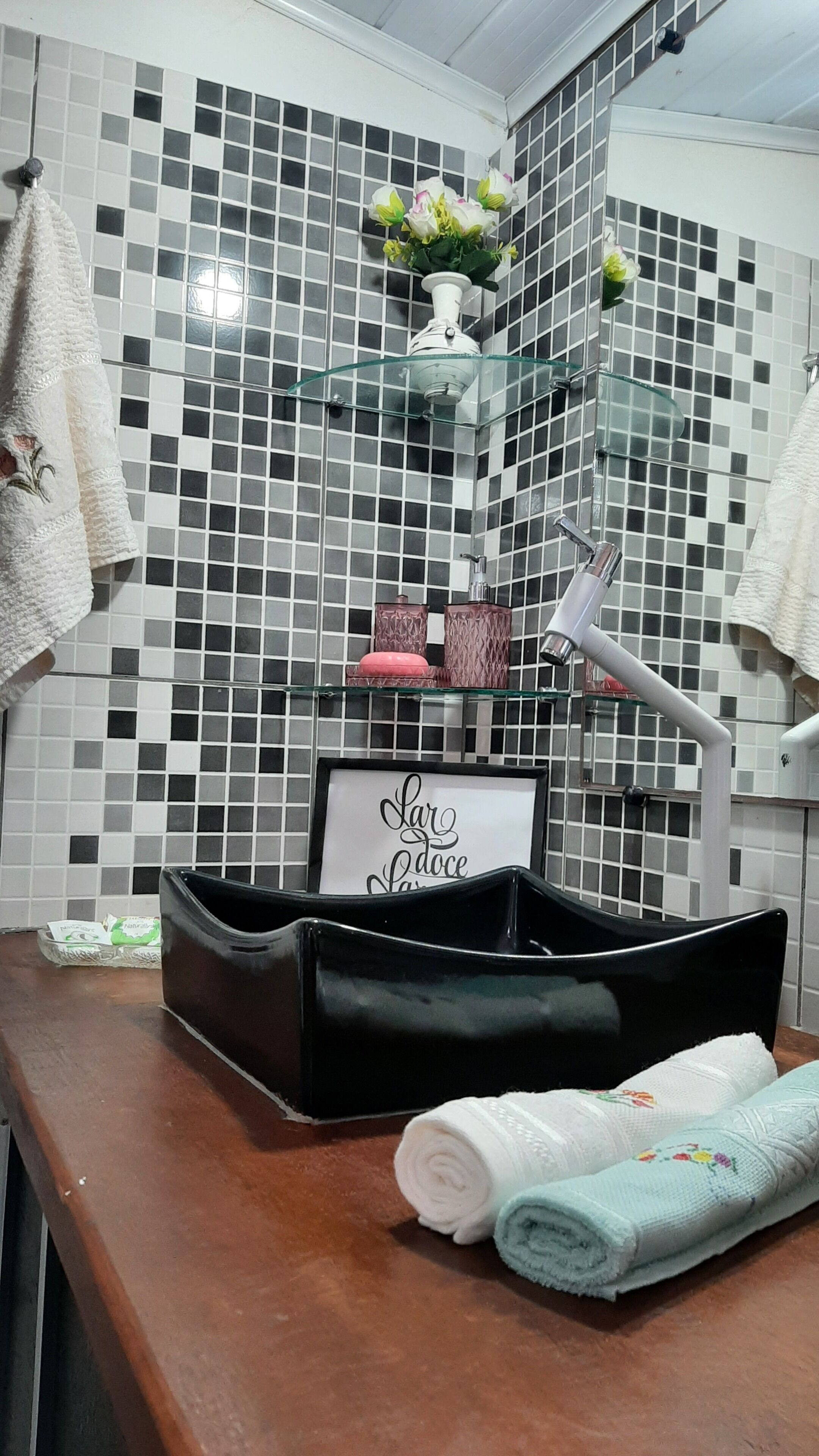 Une salle de bain avec un mur recouvert de carreaux, une étagère en verre où sont posés des articles de toilette, un panneau décoratif encadré et des serviettes sur le comptoir.