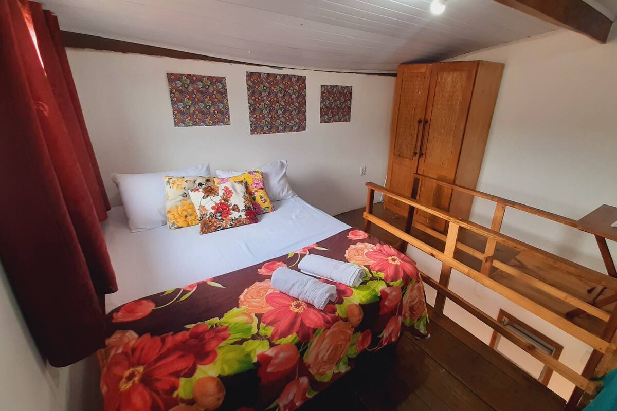 A bedroom with a bed, floral bedspread, wooden headboard, and a small table with a vase.