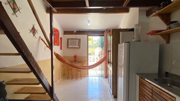 A narrow hallway with a hammock, a bed, a kitchen area with a refrigerator, and a dining area with a table and chairs. - Casas Vasconcelos (Itacaré)