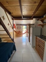 Um corredor estreito com uma rede, uma cama, uma cozinha com geladeira e um espaço de refeições com mesa e cadeiras.