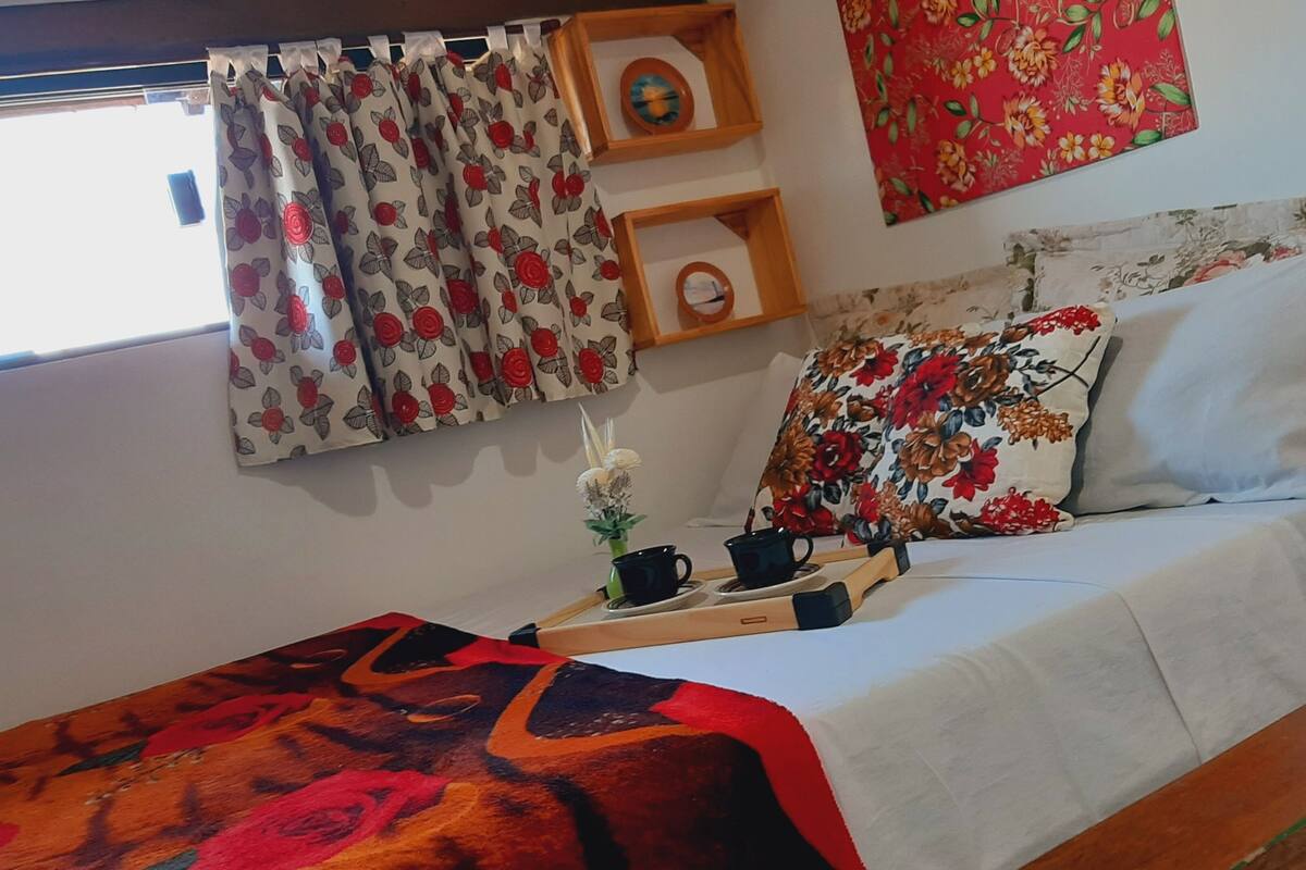 A cozy room with a bed, a small table with cups, a vase with flowers, and a window with curtains.