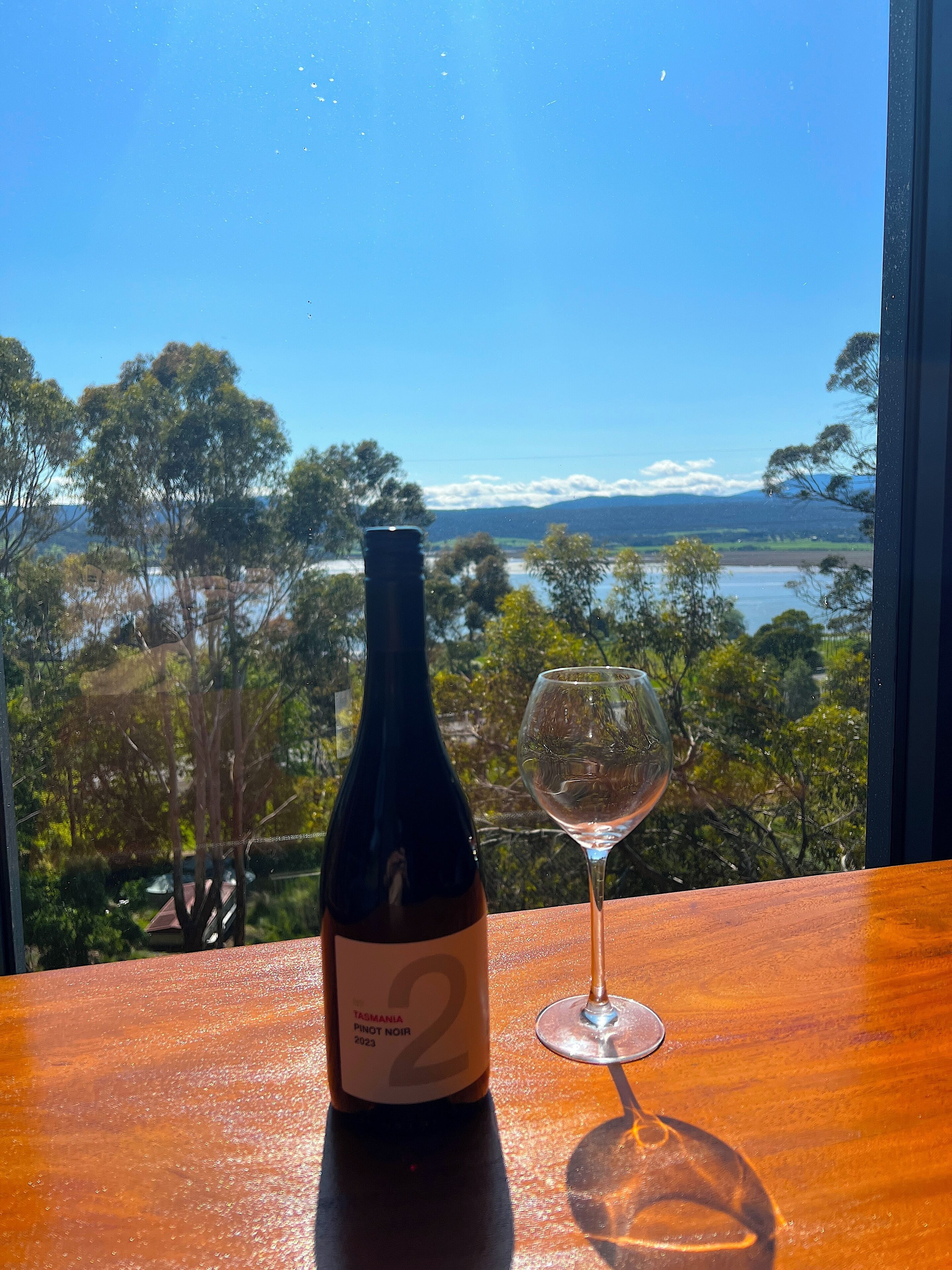 A bottle of Tasmanian Pinot Noir 2023 and a wine glass on a wooden table with a scenic view of trees and water.